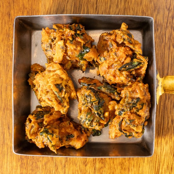 Crispy, golden-brown pakoras with visible green herbs, served in a square metal dish on a wooden table.