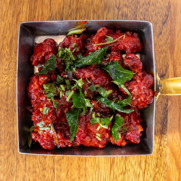 Bright red chicken 65, garnished with fresh green herbs, in a metal serving dish on a wooden table.