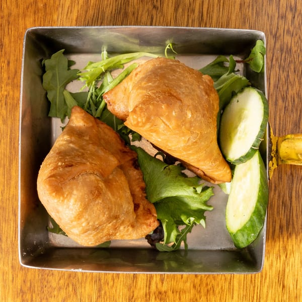 Two golden-brown samosas on a bed of green lettuce and cucumber slices in a metal tray.